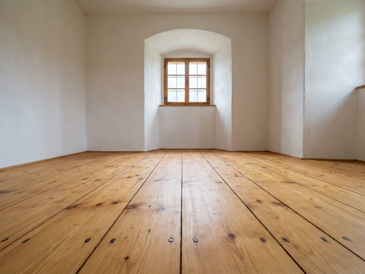 Breite, frisch geschliffene und geölte Holzdielen mit Ästen und alten Nagelköpfen in einem leeren Bauernhauszimmer mit Fensternische